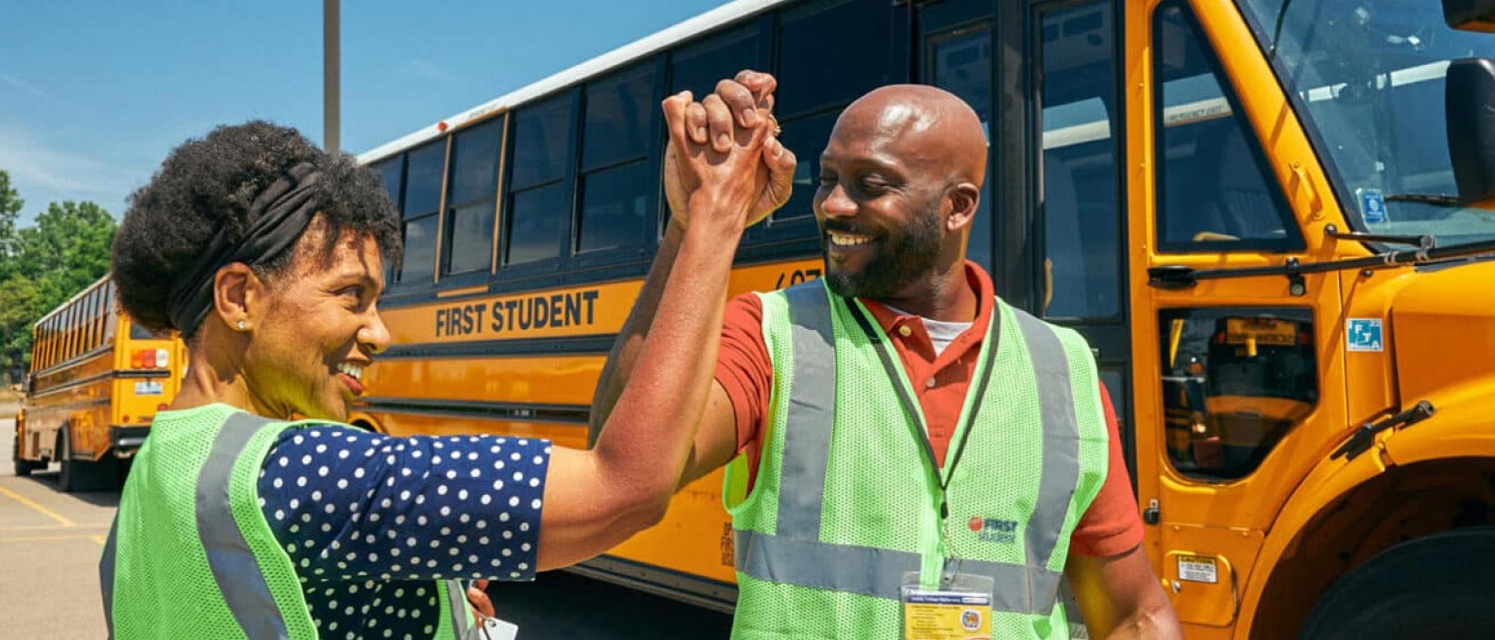 Two First Student employees giving each other a high five in front of a First Student school bus